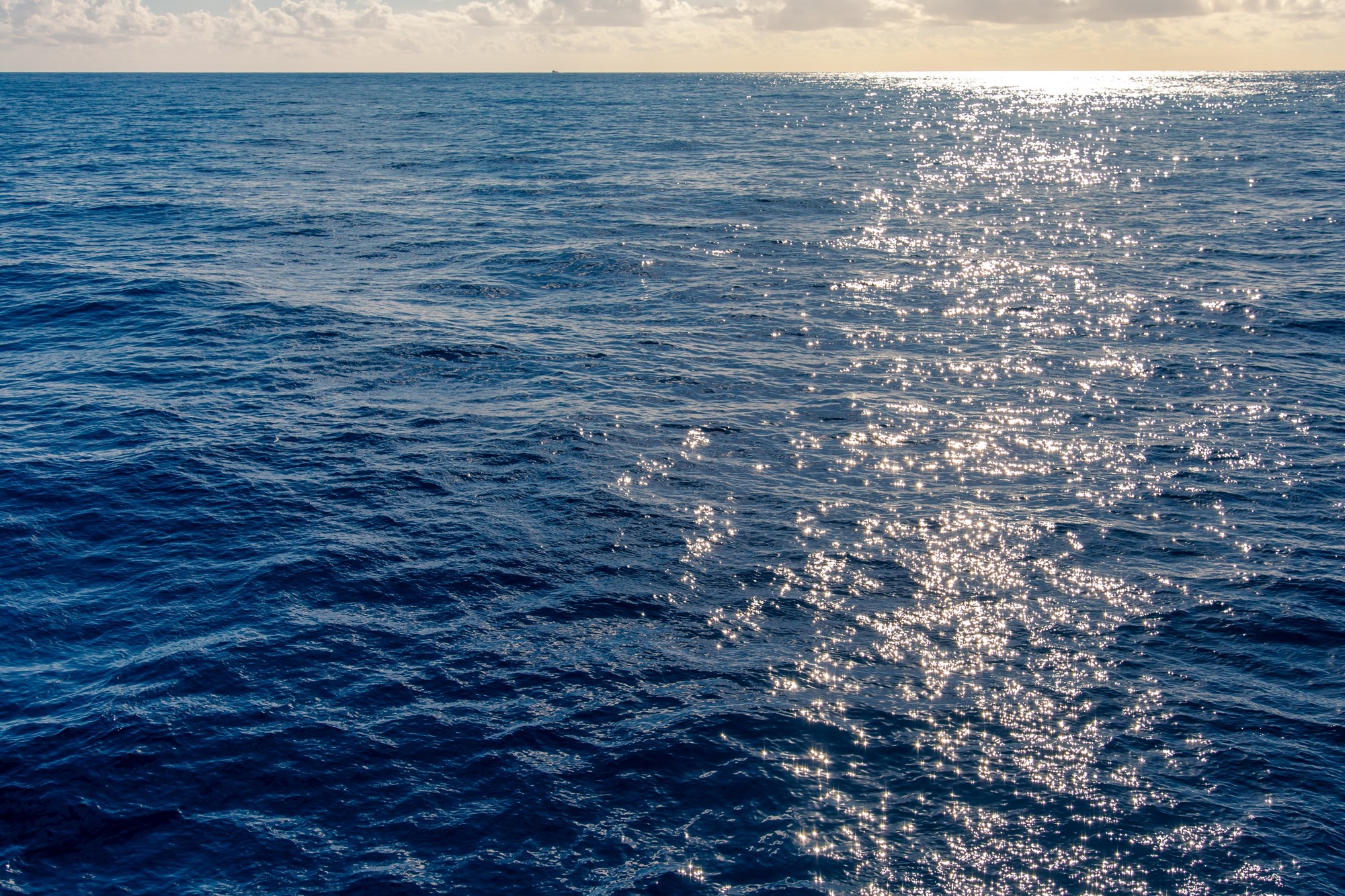 Imagem do oceano com água bem azul, aparecendo o céu com algumas nuvens ao fundo, no horizonte, com o reflexo dos raios de sol na água.