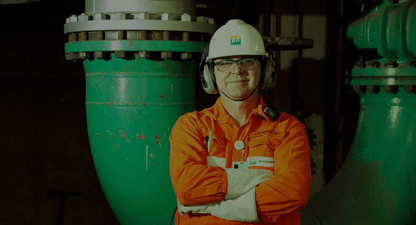 Funcionário com carreira na Petrobras, usando uniforme e equipamento de segurança, de braços cruzados.